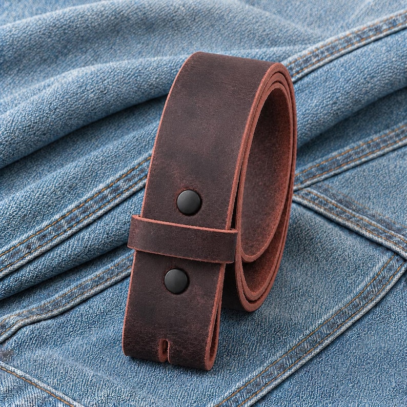May include: A rolled, dark brown leather belt with two black snap closures. The belt is made of a distressed leather material and is set against a blue denim background.