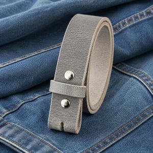 Op de afbeelding: Een opgerolde grijze leren riem met zilverkleurige klinknagels. De riem wordt getoond op een blauwe denim achtergrond. De riem heeft een eenvoudig, klassiek ontwerp.