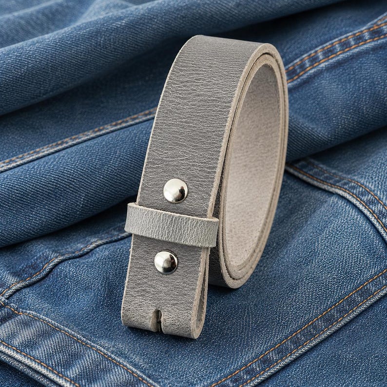Op de afbeelding: Een opgerolde grijze leren riem met zilverkleurige klinknagels. De riem wordt getoond op een blauwe denim achtergrond. De riem heeft een eenvoudig, klassiek ontwerp.