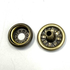 May include: Two antique brass-colored snap fasteners. One piece has a circular design with the letters "HAS" visible. The other piece has a central post and a decorative, ridged pattern. These fasteners are likely for clothing or accessories.