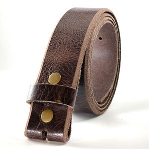 May include: A dark brown leather belt featuring a textured surface and a smooth, lighter-colored interior edge. The belt is secured with two circular, metallic brass-colored snaps located near the end, which also includes a matching leather loop.