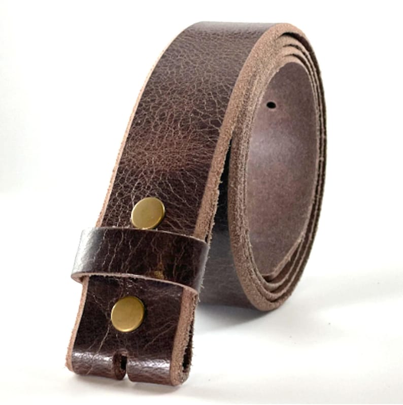 May include: A dark brown leather belt featuring a textured surface and a smooth, lighter-colored interior edge. The belt is secured with two circular, metallic brass-colored snaps located near the end, which also includes a matching leather loop.