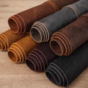 May include: A collection of rolled leather hides in various shades of brown, gray, and black. The hides are tightly rolled, showcasing the texture and color variations of the material. The leather appears to be of high quality, suitable for crafting.