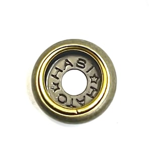 May include: A close-up of a metal button with a gold-colored inner ring and a silver-colored center. The center of the button has the word "HASIATO" stamped around the edge. The outer ring is a darker, muted gold color.