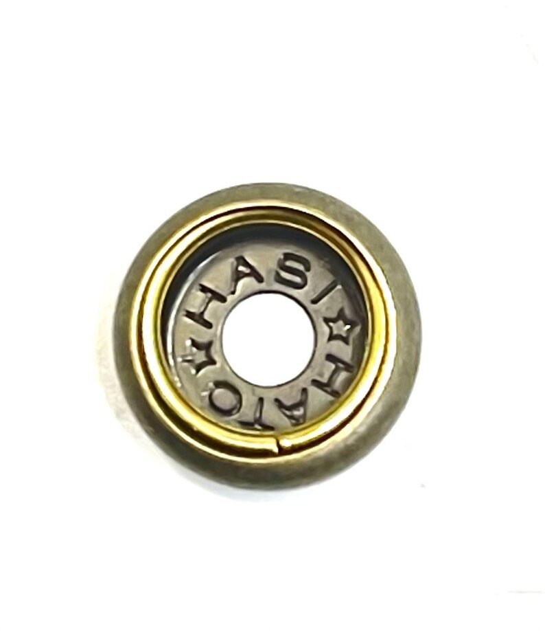 May include: A close-up of a metal button with a gold-colored inner ring and a silver-colored center. The center of the button has the word "HASIATO" stamped around the edge. The outer ring is a darker, muted gold color.