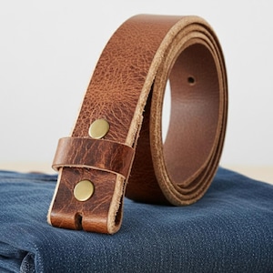 Op de afbeelding: Een opgerolde, bruine leren riem met messingkleurige klinknagels. De riem is gemaakt van getextureerd leer en lijkt ongedragen. De riem ligt op een blauw denim oppervlak.