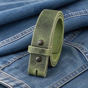 Op de afbeelding: Een opgerolde, groene leren riem met twee donkere metalen klinknagels. De riem is geplaatst tegen een achtergrond van blauwe denim stof, die de textuur en kleur benadrukt. De gesp heeft een klassiek ontwerp.