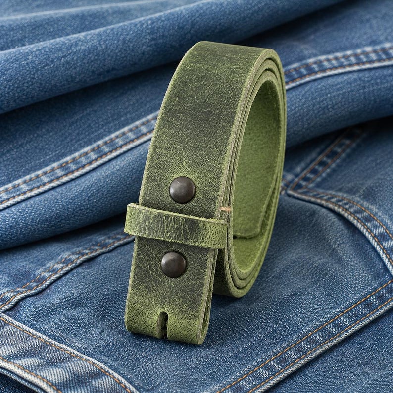 Op de afbeelding: Een opgerolde, groene leren riem met twee donkere metalen klinknagels. De riem is geplaatst tegen een achtergrond van blauwe denim stof, die de textuur en kleur benadrukt. De gesp heeft een klassiek ontwerp.