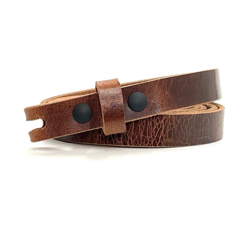 May include: A narrow, dark brown leather strap with a visible grain texture. The strap is coiled and features two matte black circular metal rivets securing the leather loop. The end of the strap has a small, U-shaped cutout notch detail.