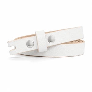 May include: A white leather strap with a textured surface, featuring two polished silver-tone metal snap buttons and a matching sliding leather loop. The strap is coiled in a circular shape, revealing a natural light beige underside.