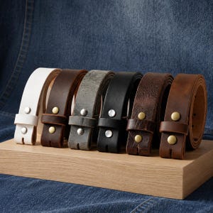 Puede incluir: Una colecci&oacute;n de correas de cintur&oacute;n de cuero en varios colores, incluyendo blanco, marr&oacute;n, gris y negro, exhibidas sobre una superficie de madera. Cada cintur&oacute;n presenta una hebilla y est&aacute; hecho de cuero de alta calidad.