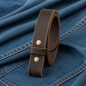 Puede incluir: Un cintur&oacute;n de cuero marr&oacute;n oscuro enrollado, con dos botones de color lat&oacute;n. El cintur&oacute;n est&aacute; desabrochado y listo para ser sujetado a un pantal&oacute;n. El cintur&oacute;n est&aacute; sobre un fondo de mezclilla azul.