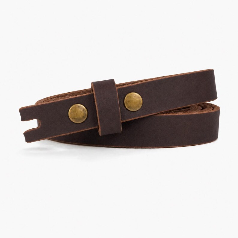 May include: A dark brown leather strap coiled into a circular shape. The strap features two circular brass-colored metal rivets and a matching leather loop. One end of the strap has a small rectangular notch cut out of the center of the edge.