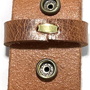 Puede incluir: Primer plano de una correa de cuero marr&oacute;n con dos cierres met&aacute;licos de presi&oacute;n y un peque&ntilde;o acento rectangular de lat&oacute;n. El cuero tiene un aspecto texturizado y desgastado, lo que sugiere un estilo vintage o r&uacute;stico. Los herrajes met&aacute;licos a&ntilde;aden contraste.