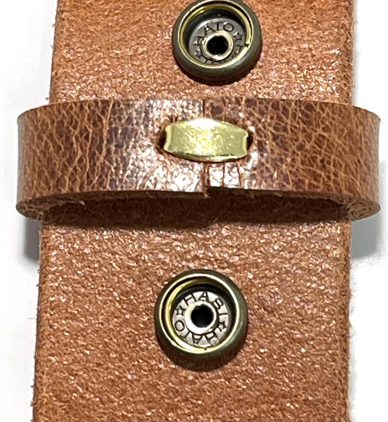 Puede incluir: Primer plano de una correa de cuero marr&oacute;n con dos cierres met&aacute;licos de presi&oacute;n y un peque&ntilde;o acento rectangular de lat&oacute;n. El cuero tiene un aspecto texturizado y desgastado, lo que sugiere un estilo vintage o r&uacute;stico. Los herrajes met&aacute;licos a&ntilde;aden contraste.