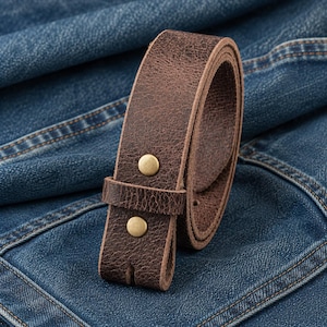 Op de afbeelding: Een opgerolde, donkerbruine leren riem met twee messingkleurige klinknagels. De riem is gemaakt van getextureerd leer en staat tegen een achtergrond van blauwe spijkerbroeken.
