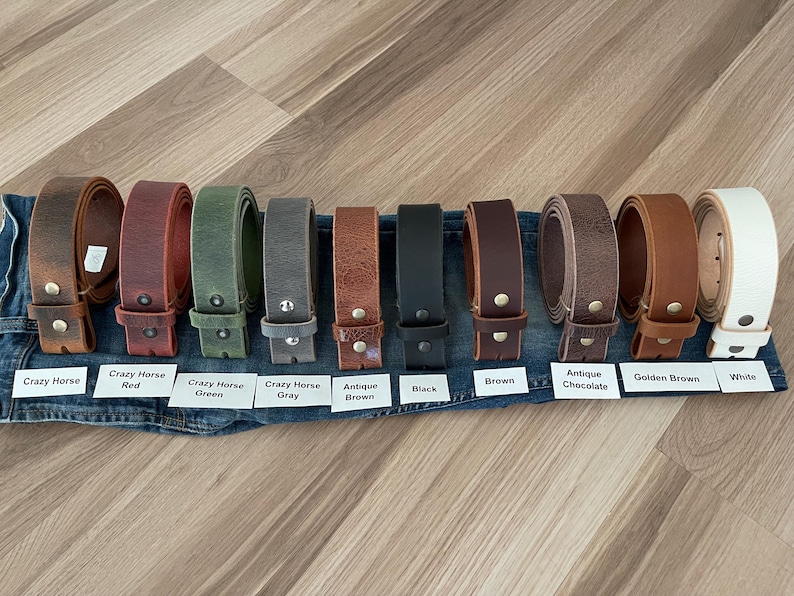 May include: A collection of leather belts in various colors, including brown, red, green, gray, and black. Each belt is displayed with a metal buckle and a label indicating the color name. The belts are arranged on a denim surface.
