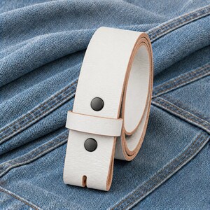 May include: A white leather belt with black rivets. The belt is rolled up, showcasing its clean design and texture. The background is a denim fabric, providing a contrasting backdrop.
