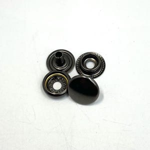 May include: Four dark gray metal snap fasteners. The snaps consist of two parts: a cap and a socket. The cap is a rounded, smooth, solid piece. The socket has a ring of small holes around the edge.