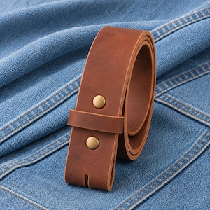 May include: A rolled brown leather belt with two brass-colored rivets and a belt loop. The belt is set against a blue denim background, showcasing its texture and craftsmanship. The belt is a classic accessory.