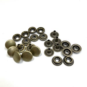 May include: A collection of antique brass-colored snap fasteners, including cap and socket components. The fasteners are arranged in groups, showcasing the different parts needed for assembly. Ideal for sewing and crafting projects.
