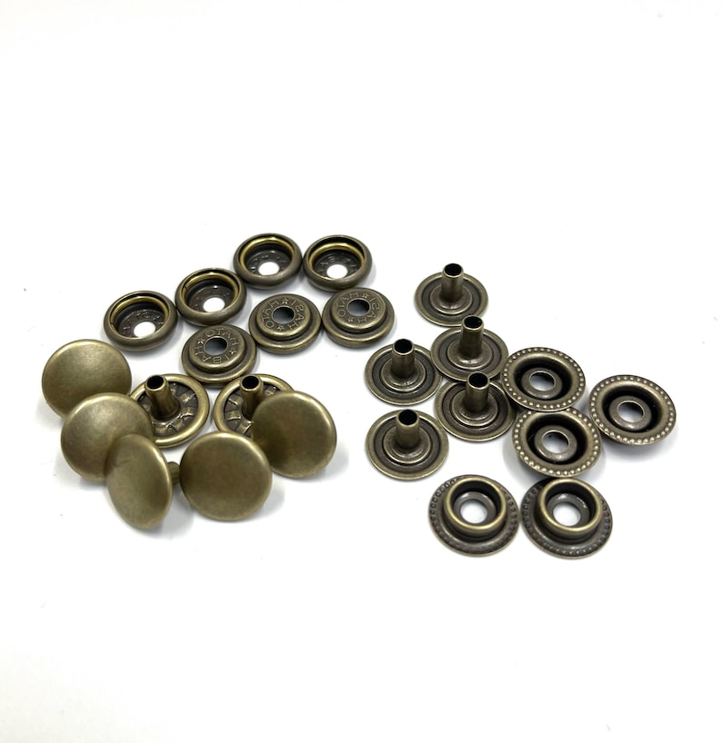 May include: A collection of antique brass-colored snap fasteners, including cap and socket components. The fasteners are arranged in groups, showcasing the different parts needed for assembly. Ideal for sewing and crafting projects.
