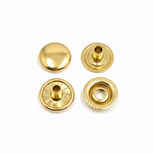 May include: Four gold-colored snap fasteners are displayed against a white background. One is a solid, rounded cap, while the others show the different parts of the snap, including the socket and stud. These fasteners are likely used for clothing or accessories.