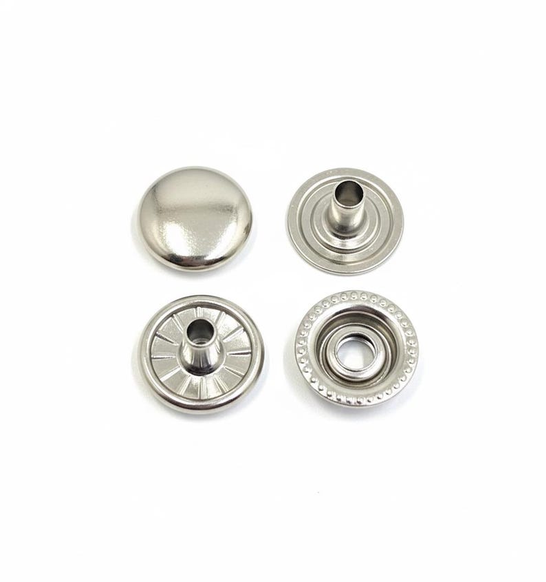 May include: Four silver-colored snap fasteners are displayed against a white background. The set includes two male and two female components. The top piece is a smooth, rounded cap. The other pieces have a central post and a circular design.
