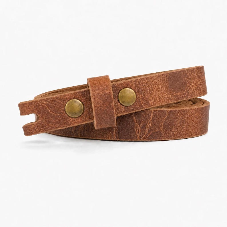 May include: A thin, brown leather strap featuring a textured surface and two circular brass-colored metal studs. The strap is coiled into a circular shape, with a small notch cut into one end and a sliding leather loop securing the overlapping section.