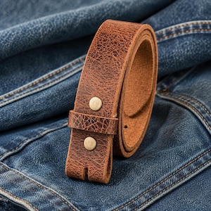 Op de afbeelding: Een opgerolde bruine leren riem met een getextureerd oppervlak en twee messingkleurige klinknagels. De riem is geplaatst tegen een achtergrond van blauwe denimstof.