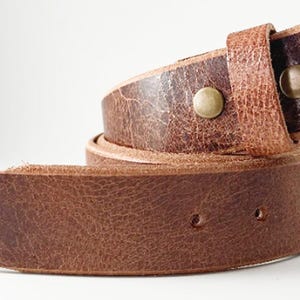 May include: A brown leather belt with a textured, distressed finish. The belt features a rounded tip, a series of punched holes for adjustment, and two metallic brass-colored snaps securing the folded end loop in a coiled position.