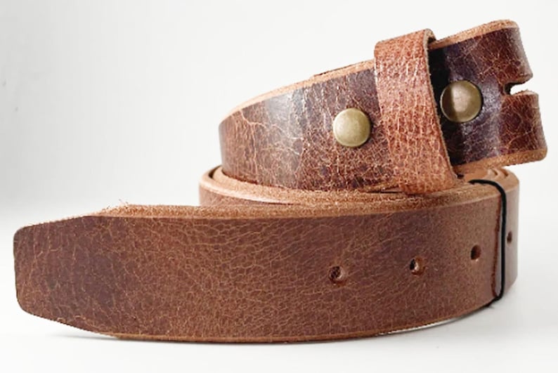 May include: A brown leather belt with a textured, distressed finish. The belt features a rounded tip, a series of punched holes for adjustment, and two metallic brass-colored snaps securing the folded end loop in a coiled position.