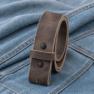May include: A rolled, brown leather belt with two black snap closures. The belt is made of textured leather and is designed to be worn with jeans or pants. The belt is shown on a blue denim background.