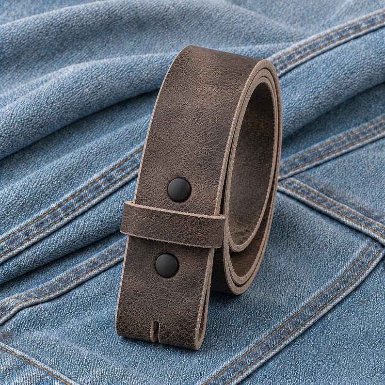 May include: A rolled, brown leather belt with two black snap closures. The belt is made of textured leather and is designed to be worn with jeans or pants. The belt is shown on a blue denim background.