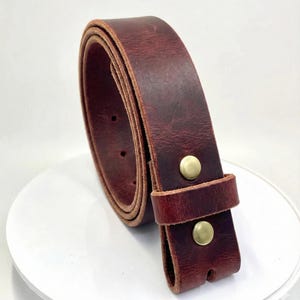 Op de afbeelding: Een opgerolde, bordeauxrode leren riem met messingkleurige klinknagels. De riem is gemaakt van dik, getextureerd leer. De riem heeft een klassiek, eenvoudig ontwerp. De riem is een veelzijdig accessoire. De riem is ongeveer 100 cm lang.