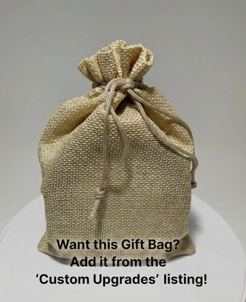 Op de afbeelding: Een beige jute cadeauzak met een trekkoordsluiting. De zak is aan de bovenkant vastgebonden en heeft de tekst "Want this Gift Bag? Add it from the 'Custom Upgrades' listing!" op de voorkant.