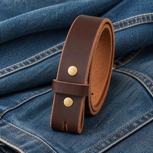Op de afbeelding: Een bruine leren riem met messingkleurige klinknagels. De riem is losgemaakt en ligt op een blauw denim oppervlak. De binnenkant van de riem is een lichtere bruintint.