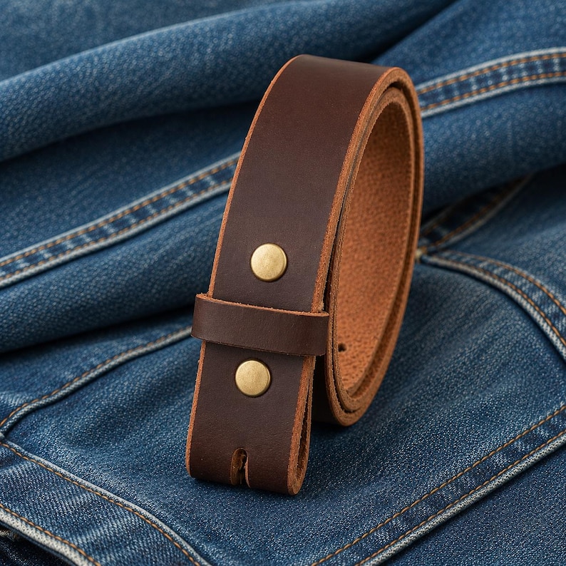 Op de afbeelding: Een bruine leren riem met messingkleurige klinknagels. De riem is losgemaakt en ligt op een blauw denim oppervlak. De binnenkant van de riem is een lichtere bruintint.