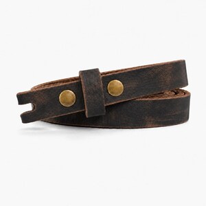 May include: A dark brown leather strap with a distressed finish, featuring two circular brass-toned metal studs and a matching leather loop. The strap is coiled, showing a notched end and a smooth, flat texture throughout the material.