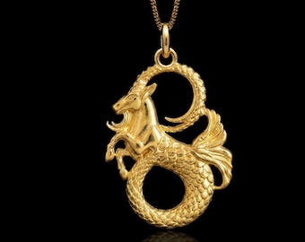 May include: A gold-toned necklace featuring a pendant shaped like a Capricorn sea-goat. The pendant displays a detailed goat head with curved horns and a scaled, fish-like tail curled into a circular form, suspended from a delicate gold chain.