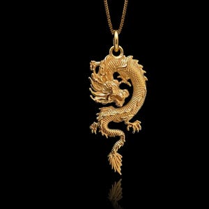 May include: A gold dragon pendant necklace. The dragon is highly detailed with scales, claws, and a flowing mane. The pendant hangs from a fine chain against a black backdrop.