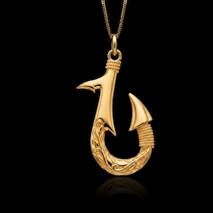 May include: A gold-colored pendant shaped like a fish hook suspended from a matching gold-colored chain. The pendant features intricate scrollwork engravings along the curved base and textured rope-like detailing at the top and bottom points.