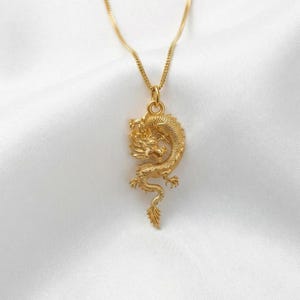May include: A gold pendant necklace with a detailed dragon design. The dragon is intricately sculpted with scales, a flowing mane, and a curled tail. The necklace hangs on a delicate chain, set against a white background.