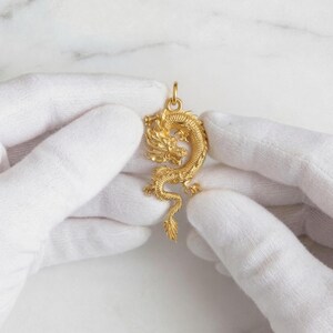 May include: A gold dragon pendant with detailed features, held by gloved hands. The dragon is in a curled pose, with a detailed head, body, and tail. The pendant has a loop at the top for a chain.