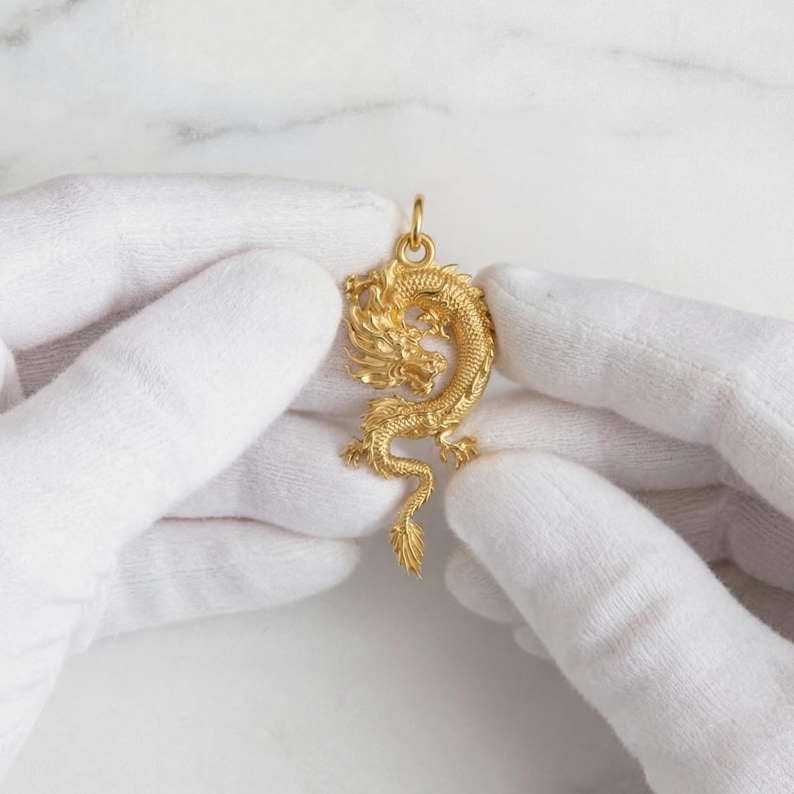 May include: A gold dragon pendant with detailed features, held by gloved hands. The dragon is in a curled pose, with a detailed head, body, and tail. The pendant has a loop at the top for a chain.