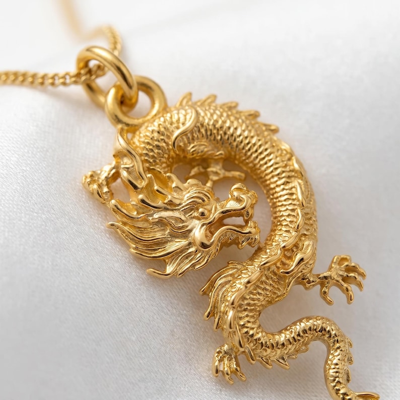May include: A gold dragon pendant on a necklace. The dragon is highly detailed with scales, claws, and a flowing mane. The pendant is suspended from a fine gold chain, creating a striking piece of jewellery.