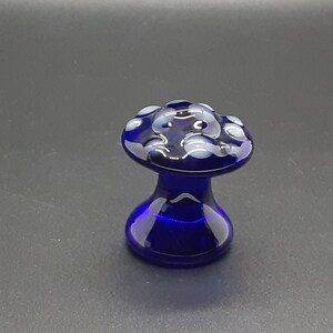 Op de afbeelding: Een kleine blauwe glazen paddenstoel met witte stippen. De paddenstoel is een decoratief object.