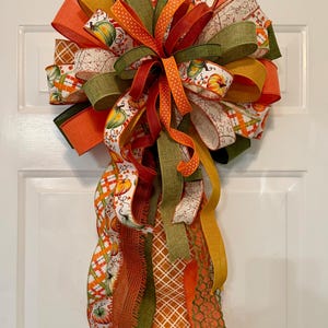 May include: A decorative door bow featuring a mix of wired ribbons in orange, olive green, and white. The ribbons display patterns of pumpkins, autumn leaves, and plaid designs. The bow has a large, full top section with long, cascading tails.