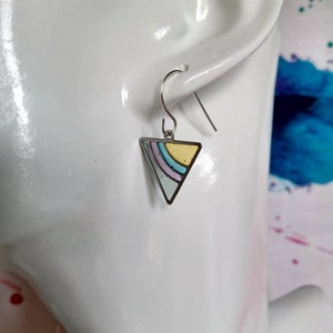 Op de afbeelding: Zilveren driehoekige oorbellen met een pastelkleurig regenboogontwerp. De oorbellen zitten op een wit mannequinhoofd.