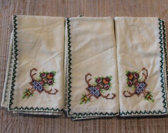 Puede incluir: Tres servilletas de tela color crema con diseños florales bordados. Cada servilleta presenta un patrón de punto de cruz de flores en tonos rojos, naranjas, verdes y azules. Los bordes están acabados con una puntada en zigzag verde.